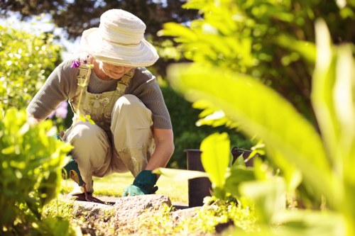 A woman wearing a wide-brimmed hat, light-colored trousers, a grey top, and gardening gloves is working in a well-maintained backyard garden in Stratford. She is kneeling on the grass and appears to be planting or tending to a flower bed bordered by small, evenly spaced bricks or stones, with rich soil visible within the bed. The surrounding garden includes a lush, green lawn with dense foliage, shrubs, and trees in the background, some of which have bright green leaves reflecting sunlight. Paving or decking areas are visible at the edges of the garden, and the scene suggests a warm, sunny day with soft natural light illuminating the garden space. The overall environment is tidy and inviting, characteristic of outdoor spaces maintained by local gardening services in Stratford, with a focus on lush plantings and well-organised garden features.